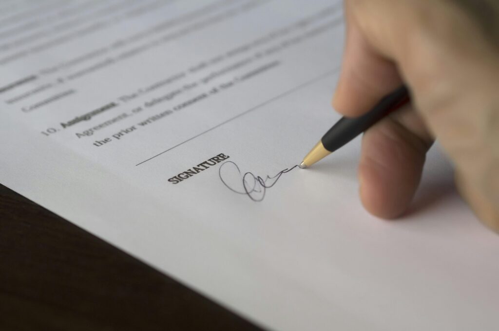 A hand signs a formal contract with a pen on a wooden desk.